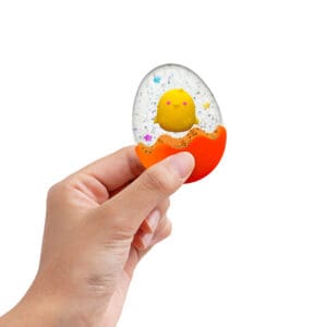 SQTEND25_Image5 A hand holds a transparent egg-shaped object with a small yellow chick inside. The bottom half of the egg is orange, resembling a cracked shell. Glitter and colorful stars decorate the inside, adding a playful touch. The background is plain white.